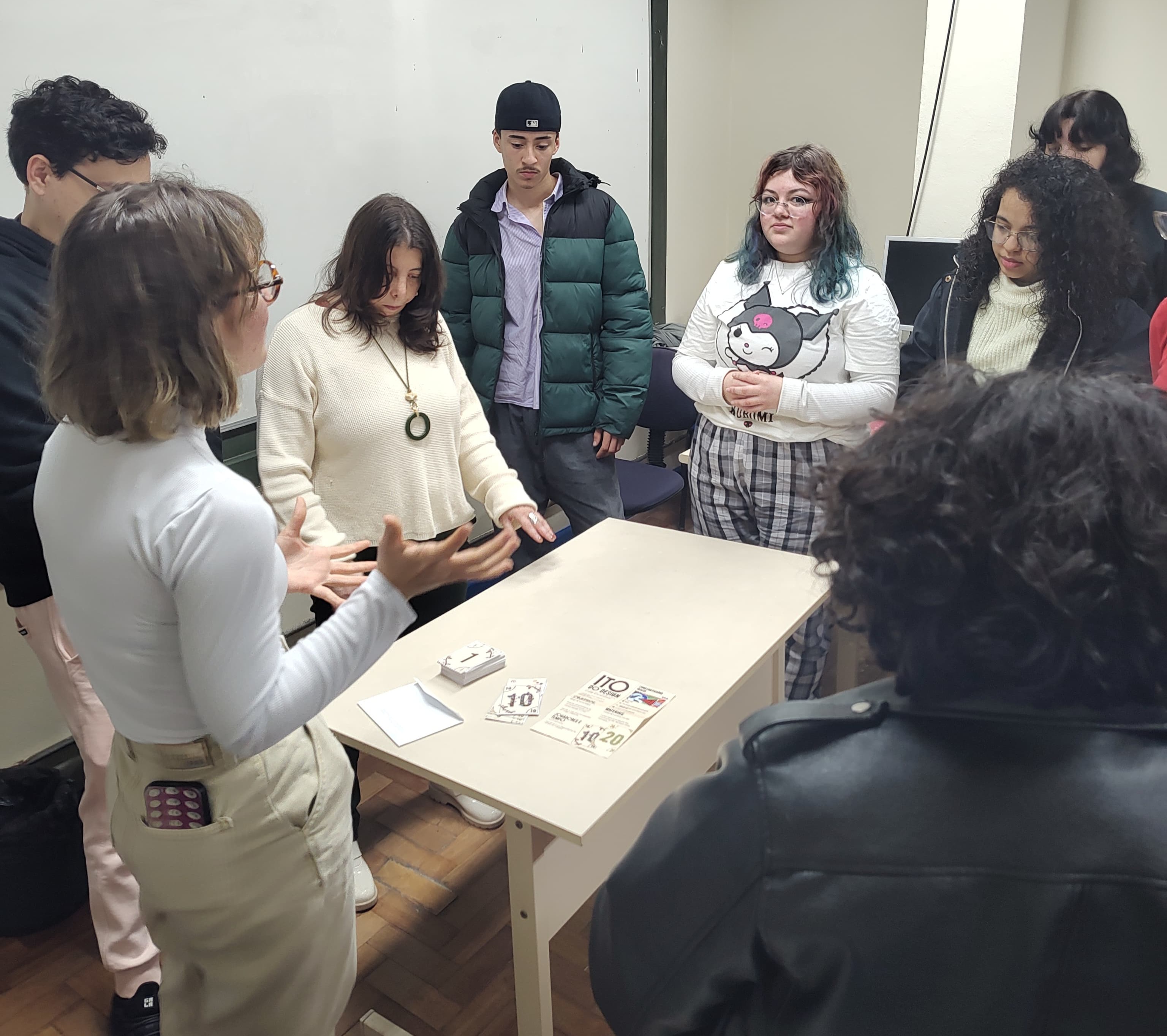 Na fotografia de Anna Carolina Batista, a professora da UFPel Chris Ramil, mulher branca e de cabelos longos e escuros, vestindo blusa de cor branca e um colar com adereço circular na ponta, aparece em pé, em frente a uma mesa, rodeada por oito estudantes, durante aula do curso de Design. A professora, com a cabeça baixa, olha para jogo de cartas disposto em cima de mesa, enquanto uma estudante da UFPel aparece de perfil, mulher branca, com cabelos curtos e claros, vestindo roupas claras e utilizando óculos de lentes arredondadas, apresenta o trabalho realizado na disciplina, movimentando as duas mãos enquanto fala. 